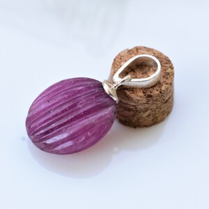 Puede incluir: Un colgante ovalado de color morado con un aro de plata. El colgante tiene una textura estriada y está colocado sobre un fondo de corcho.
