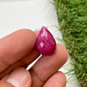 May include: A faceted, teardrop-shaped ruby gemstone with a wire loop for jewelry making. The ruby is a deep, vibrant red color and has a polished, reflective surface. The gemstone is held between fingers against a white and green background.