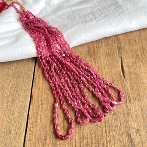May include: A strand of pink oval-shaped beads, strung on a gold thread. The beads are arranged in a cascading pattern, with the ends of the strand hanging down.
