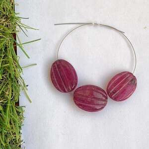 May include: Three oval-shaped, pink gemstone beads with a carved, striped pattern. The beads are strung on a silver wire.