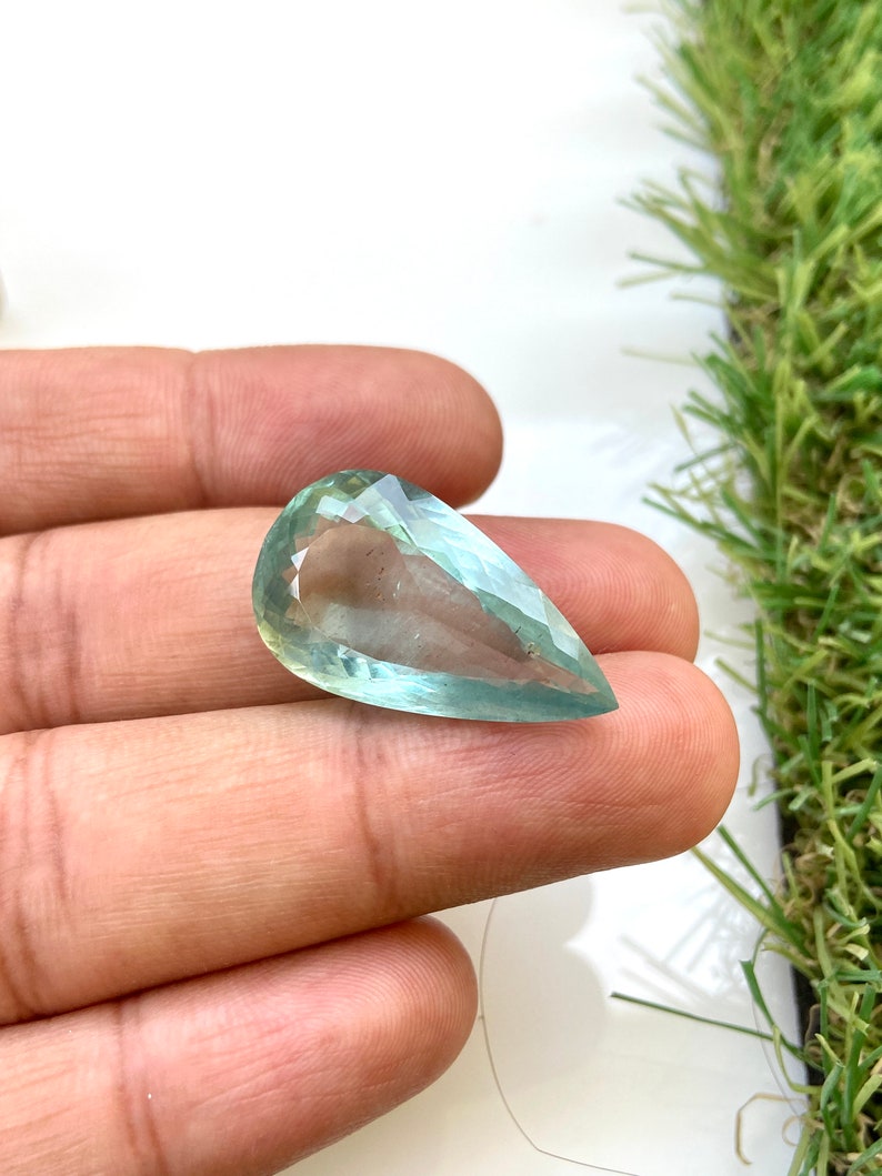 May include: A teardrop-shaped aquamarine gemstone with a light blue-green color. The gemstone is faceted and has a smooth, polished surface.