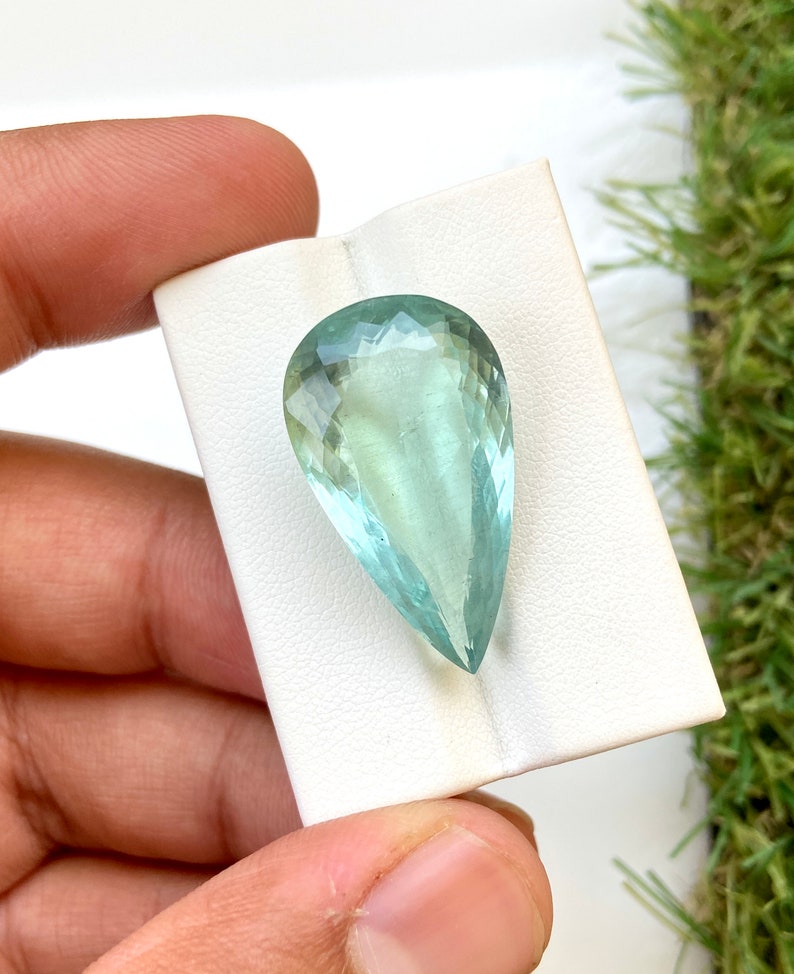 May include: A pear-shaped aquamarine gemstone with a light blue color. The gemstone is faceted and has a smooth, polished finish.