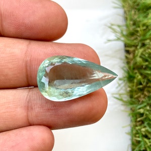 May include: A teardrop-shaped aquamarine gemstone with a light blue-green color. The gemstone is faceted and has a smooth, polished finish.