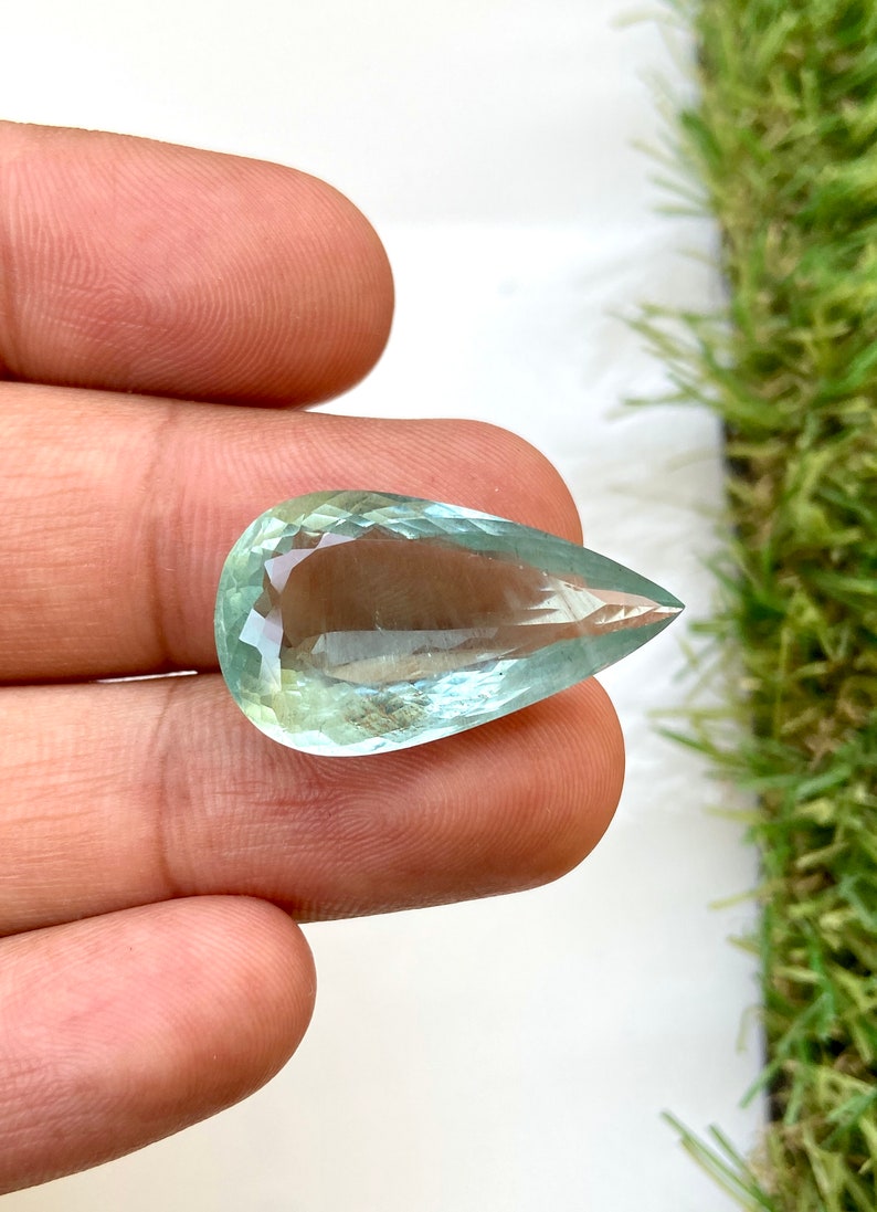 May include: A teardrop-shaped aquamarine gemstone with a light blue-green color. The gemstone is faceted and has a smooth, polished finish.