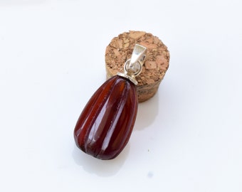 May include: A dark red, faceted, teardrop-shaped pendant with a silver bail. The pendant is sitting on a cork.