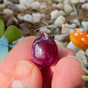 May include: A deep red, teardrop-shaped gemstone pendant with a silver wire and gold accent. The polished stone is held by a silver wire loop. The background features small white and tan stones and a miniature orange mushroom.