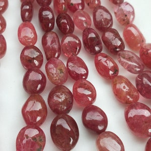 Peut inclure: Gros plan sur des brins de pierres précieuses polies, de forme ovale, dans des tons de rouge et de rose. Les pierres ont une surface lisse et brillante et sont enfilées, suggérant qu'elles sont destinées à la création de bijoux. Les pierres sont sur fond blanc.