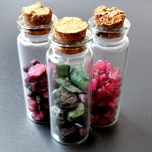 Puede incluir: Tres pequeñas botellas de vidrio con tapones de corcho, cada una llena de piedras preciosas de diferentes colores. La botella de la izquierda contiene piedras preciosas rojas, la botella del medio contiene piedras preciosas verdes y la botella de la derecha contiene piedras preciosas rojas.