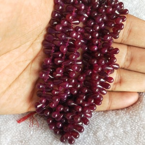 Op de afbeelding: Een streng dieprode edelsteen kralen in een druppelvorm. De kralen zijn geregen aan een dun draad en zijn gerangschikt in een cascade patroon.