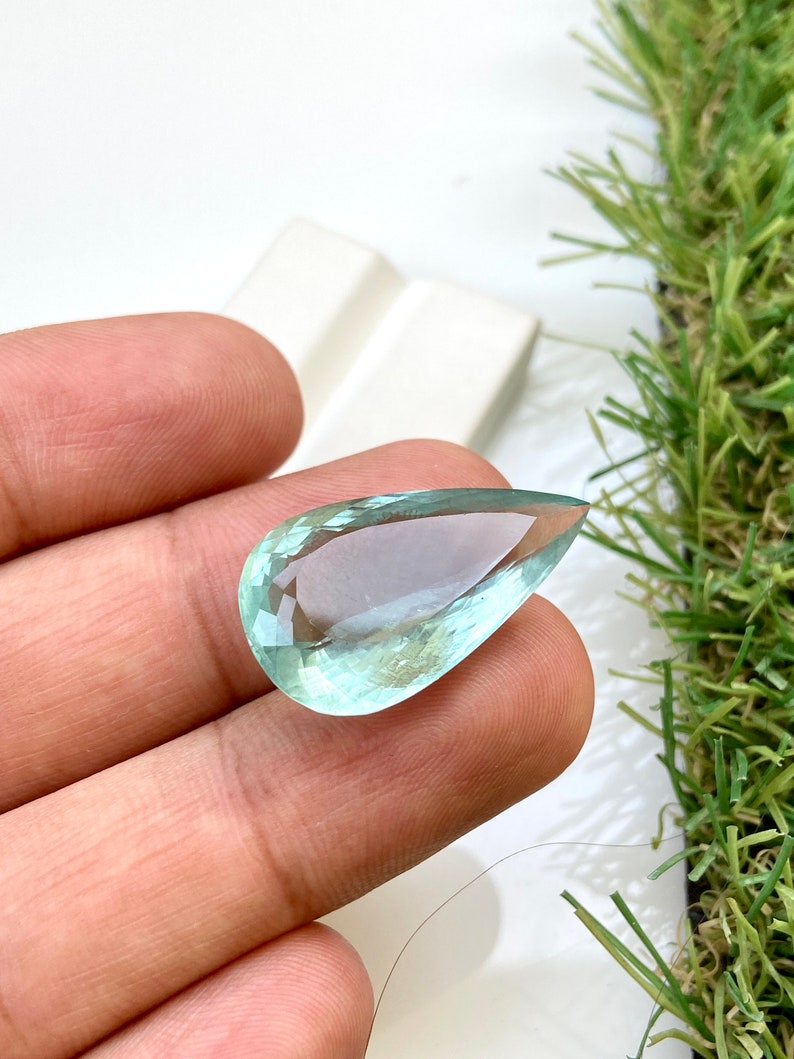 May include: A teardrop-shaped aquamarine gemstone with a light blue color. The gemstone is faceted and has a high degree of clarity.