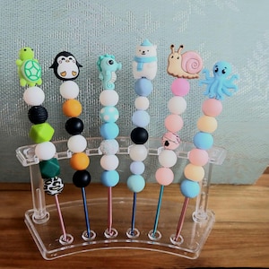 Può includere: Sei uncinetti in metallo con manici in perline di silicone, ognuno sormontato da una diversa figura animale tra cui tartaruga, pinguino, cavalluccio marino, orso polare, lumaca e polpo. Le perline hanno vari colori e motivi su un supporto.