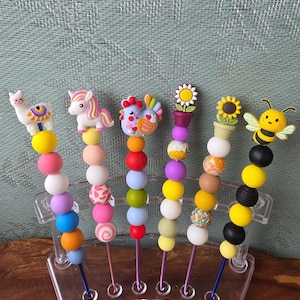 May include: Six decorative pens featuring silicone character toppers including a llama, unicorn, chicken, potted flower, sunflower, and bee. Each pen is adorned with a vertical stack of colorful, round beads in various solid and patterned designs.