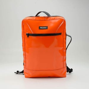 May include: Bright orange backpack with a glossy finish, featuring a black zipper and a small rectangular label that reads "Mustaches". The backpack has a top handle and adjustable straps, ideal for carrying essentials.