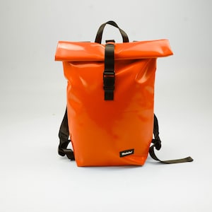 May include: A bright orange roll-top backpack with a glossy, water-resistant finish. It features a black vertical strap with a plastic buckle closure, a top handle, adjustable black shoulder straps, and a small black label reading Moustache.
