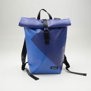 Könnte beinhalten: Ein blauer Rucksack mit Rollverschluss und geometrischem Design. Der Rucksack hat ein dunkelblaues Dreieck auf hellblauem Hintergrund, schwarze Riemen und einen schwarzen Schnallenverschluss. Der Markenname "Mustaches" ist auf der Vorderseite aufgedruckt.