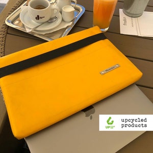 Peut inclure: Une housse d'ordinateur portable jaune avec une sangle noire et une étiquette de nom argentée qui dit "Mustache". La housse est posée sur un ordinateur portable argenté avec un logo Apple. L'ordinateur portable est sur une table en bois avec un verre de jus d'orange, une tasse de café et une plante en pot.
