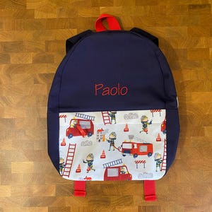 May include: A navy blue backpack featuring a red top handle and red straps. The name Paolo is embroidered in red thread on the upper section. The front pocket displays a white fabric pattern with illustrations of red fire trucks, firefighters, ladders, and orange traffic cones.