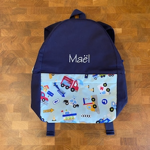 May include: A navy blue backpack featuring the name Maël embroidered in white thread on the upper section. The front pocket displays a light blue fabric pattern with various colorful vehicles including fire trucks, police cars, and boats.