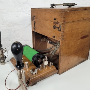Puede incluir: Una caja de madera vintage con una bobina verde y dos pomos negros. La caja tiene una tapa con bisagras y un muelle metálico en el interior. La caja está sobre una superficie blanca.