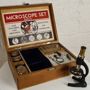 Pode incluir: Uma caixa de madeira com um conjunto de microscópio dentro. A caixa é etiquetada como 'Microscope Set Einco' e tem um selo à prova de pó. O conjunto inclui um microscópio, lâminas e um frasco para espécimes. A caixa está aberta e o microscópio é visível.