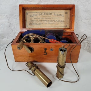 Puede incluir: Una caja de madera antigua con una manivela de latón y dos tubos de latón. La caja está etiquetada "Magneto-Electric Machine for Nervous & Other Diseases".