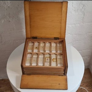 Puede incluir: Una caja de madera con tapa abatible, que contiene una colección de portaobjetos de microscopio de vidrio. La caja está dividida en compartimentos, cada uno de los cuales contiene un portaobjetos. Los portaobjetos están etiquetados con texto escrito a mano.