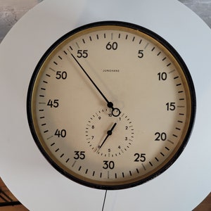 May include: A black and gold analog clock with a white face and black hands. The clock has a smaller clock face inside the larger one. The larger clock face has numbers from 5 to 60, and the smaller clock face has numbers from 1 to 10. The clock is made by Junghans.