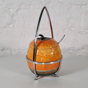 May include: Orange and black ceramic sugar bowl with a metal handle and stand. The bowl is decorated with a striped pattern.