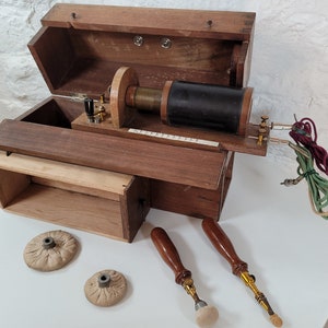 Puede incluir: Caja de madera antigua que contiene un dispositivo médico vintage con una bobina, cables y dos sondas. La caja tiene una tapa con bisagras y un pequeño cajón. El dispositivo se utiliza para tratamientos médicos.