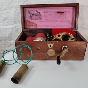 Puede incluir: Caja de madera antigua que contiene una máquina magnetoeléctrica para enfermedades nerviosas. La caja está abierta, revelando los componentes de la máquina en latón y una almohadilla de fieltro rojo. Dos electrodos de latón con cables verdes son visibles fuera de la caja.
