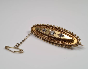 Könnte beinhalten: Eine ovale Brosche aus Gold mit einer Kette und einer Sicherheitsnadel. Die Brosche ist mit einem Perlenrand und drei klaren Steinen verziert.