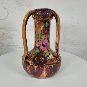 Puede incluir: Un jarrón de cerámica marrón con dos asas arqueadas y diseños florales. El jarrón presenta flores pintadas de color púrpura, rosa y verde. La base tiene un patrón geométrico en rojo y marrón. El jarrón mide unos 25 cm de alto.
