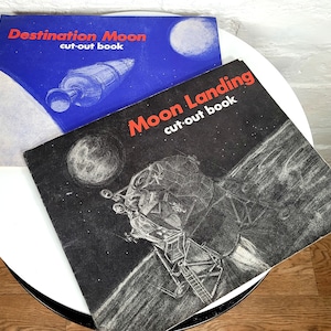 May include: Two vintage children's cut-out books about space exploration. The top book is blue with the title "Destination Moon cut-out book". The bottom book is black with the title "Moon Landing cut-out book".