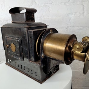May include: Antique brass and black metal slide projector with a lens and a chimney. The projector has a handle on the side and a small door on the front.