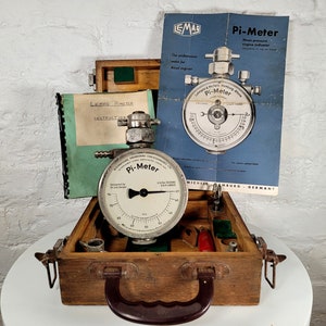 Peut inclure: Une boîte en bois vintage contenant un indicateur de moteur LeMas Pi-Meter. La boîte a une poignée en laiton et un couvercle à charnières. Le Pi-Meter est un manomètre rond avec un cadran blanc et des marquages noirs. Le manomètre est relié à un tuyau métallique avec une valve. La boîte contient également un petit livret d'instructions.