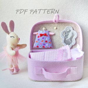 May include: A pink felt suitcase with a handle, a white felt bed with a pink and white checked blanket, a purple dress with a red bow, and a silver mirror inside. A white felt bunny wearing a pink tutu stands next to the suitcase. The text "PDF PATTERN" is written above the suitcase.