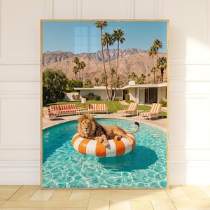 May include: A framed wall art print depicting a lion resting on a large orange and white striped circular pool float in a turquoise swimming pool. The scene features a mid-century modern house, palm trees, and mountains under a clear blue sky.