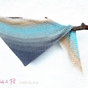Könnte beinhalten: Ein dreieckiger Häkel-Schal in Beige-, Blau- und Grautönen, der über einen Ast gelegt ist. Der Schal hat ein Farbverlaufsmuster und ist mit einer feinen Masche gearbeitet. Der Text "Stick & Fit made by Anja" ist in der linken unteren Ecke des Bildes zu sehen.
