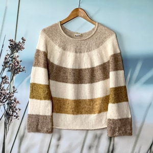 Könnte beinhalten: Ein gestreifter Pullover mit braunen, weißen und goldenen Streifen. Der Pullover hat einen runden Ausschnitt und lange Ärmel.