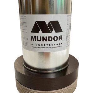 Op de afbeelding: Een zilveren verfblik met de merknaam "MUNDOR" in zwarte letters en een zwart logo. Het blik staat op een donkerbruine houten basis, die op een lichtere bruine houten basis staat.