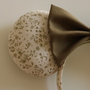 May include: A headband with two round ears and a large bow. The ears are covered in a cream-colored fabric with a floral pattern in shades of green. The bow is a solid olive green.