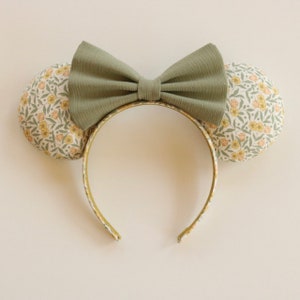 May include: A pair of green and white floral mouse ears with a green bow. The ears are made of fabric and have a floral pattern. The bow is made of a soft, textured fabric.