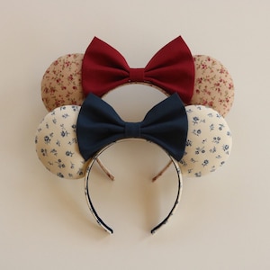 May include: A pair of mouse ears with a floral print fabric. One ear has a burgundy bow and the other has a navy blue bow.