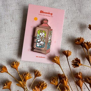 Peut inclure: Une carte rose avec le mot "Blooming" et une &eacute;pingle en &eacute;mail en forme de lanterne. L'&eacute;pingle pr&eacute;sente un motif de coton &agrave; l'int&eacute;rieur d'une lanterne avec un cadre dor&eacute;. Des fleurs oranges s&eacute;ch&eacute;es sont &eacute;parpill&eacute;es autour de la carte.