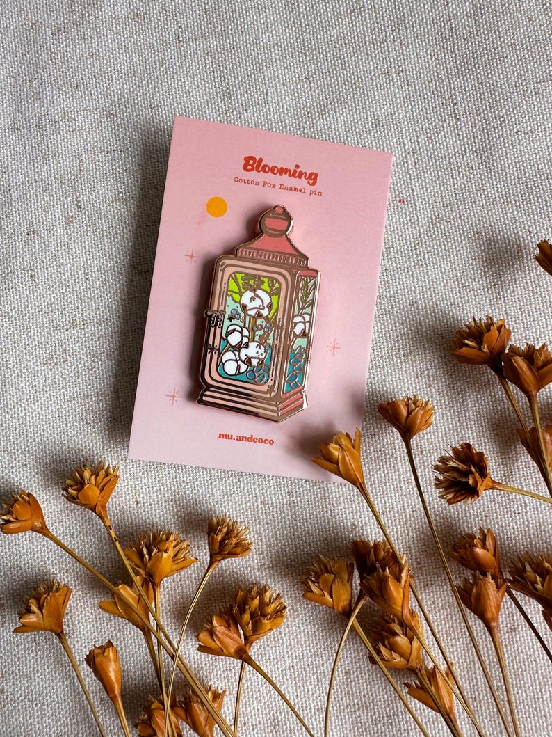 Peut inclure: Une carte rose avec le mot "Blooming" et une &eacute;pingle en &eacute;mail en forme de lanterne. L'&eacute;pingle pr&eacute;sente un motif de coton &agrave; l'int&eacute;rieur d'une lanterne avec un cadre dor&eacute;. Des fleurs oranges s&eacute;ch&eacute;es sont &eacute;parpill&eacute;es autour de la carte.