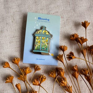 Peut inclure: &Eacute;pinglette en &eacute;mail "Blooming" avec un motif floral jaune et bleu dans une cage d&eacute;corative. L'&eacute;pinglette est pos&eacute;e sur une carte bleue avec le mot "Blooming" et le texte "bunny bud enamel pin". Des fleurs orange s&eacute;ch&eacute;es sont &eacute;parpill&eacute;es.