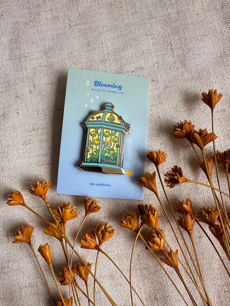 Peut inclure: &Eacute;pinglette en &eacute;mail "Blooming" avec un motif floral jaune et bleu dans une cage d&eacute;corative. L'&eacute;pinglette est pos&eacute;e sur une carte bleue avec le mot "Blooming" et le texte "bunny bud enamel pin". Des fleurs orange s&eacute;ch&eacute;es sont &eacute;parpill&eacute;es.