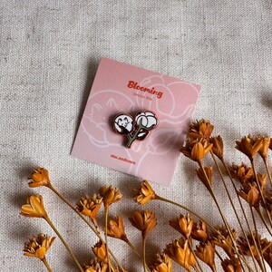 Peut inclure: Une &eacute;pingle en coton avec un motif de plante de coton, pos&eacute;e sur une carte rose avec le texte "Blooming Cotton Fox". L'&eacute;pingle est noire, blanche et verte. Des fleurs oranges s&eacute;ch&eacute;es sont &eacute;parpill&eacute;es autour de la carte.