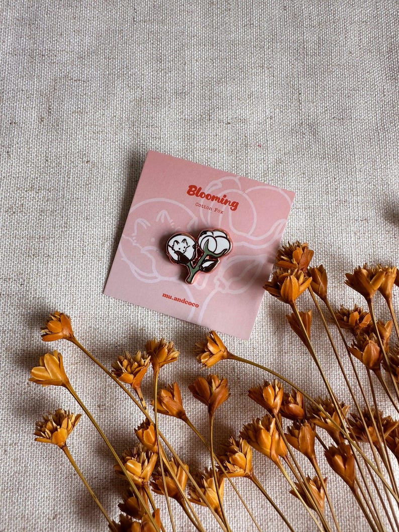 Peut inclure: Une &eacute;pingle en coton avec un motif de plante de coton, pos&eacute;e sur une carte rose avec le texte "Blooming Cotton Fox". L'&eacute;pingle est noire, blanche et verte. Des fleurs oranges s&eacute;ch&eacute;es sont &eacute;parpill&eacute;es autour de la carte.