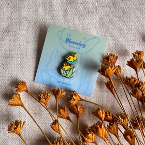 Peut inclure: Une carte bleu clair avec le texte "Blooming Bunny Bud" et une &eacute;pingle lapin. L'&eacute;pingle repr&eacute;sente un lapin jaune avec un masque bleu. Des fleurs orange s&eacute;ch&eacute;es sont dispos&eacute;es autour de la carte.